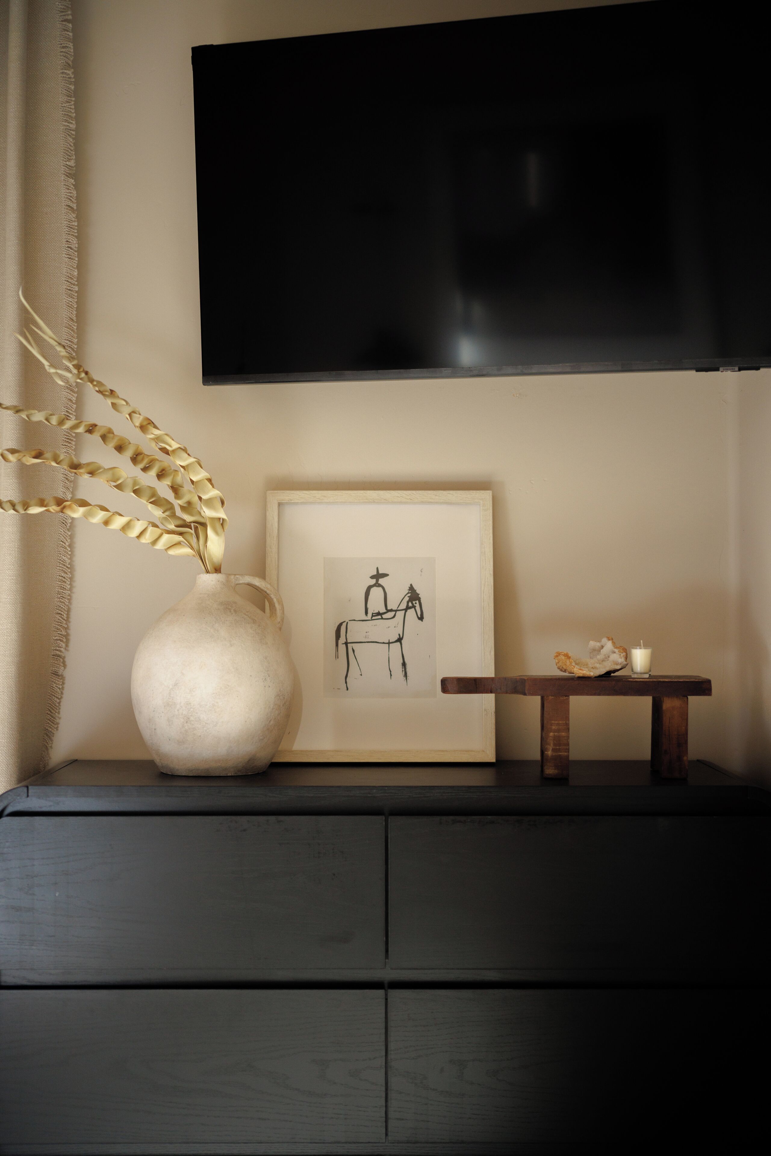 Un téléviseur fixé au mur, au-dessus d’une commode en bois foncé, sur laquelle sont posés un vase et un tableau encadré.