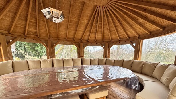 A wooden dining area with a large round table, cushioned seating, and a wooden ceiling structure. - Renovated, spacious and comfortable 26-bed mill (Saint-Ignat)