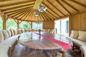 Une grande table à manger en bois, surmontée d’un plateau en verre, entourée de sièges blancs rembourrés, sous un plafond en bois agrémenté d’un lustre.