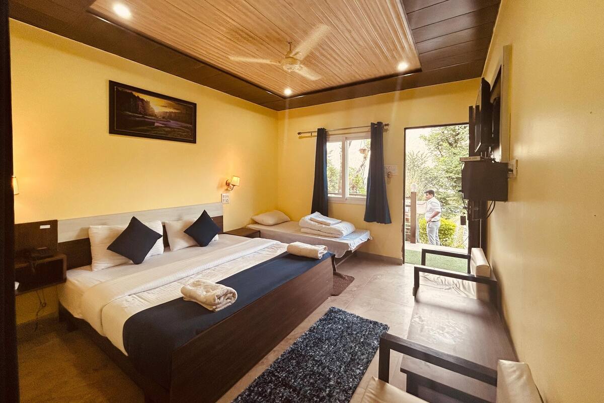 Habitación de hotel con dos camas, ventilador de techo, un cuadro en la pared, una ventana con cortinas y un televisor montado.
