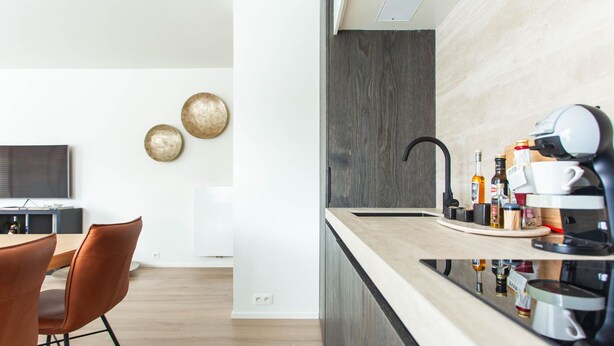A modern kitchen with a dark wood backsplash, a stainless steel sink, and a coffee machine. There are two round wall decorations and a dining area with a wooden table and chairs. - Elegant Coastal Retreat in Knokke-heist (Knokke-Heist)