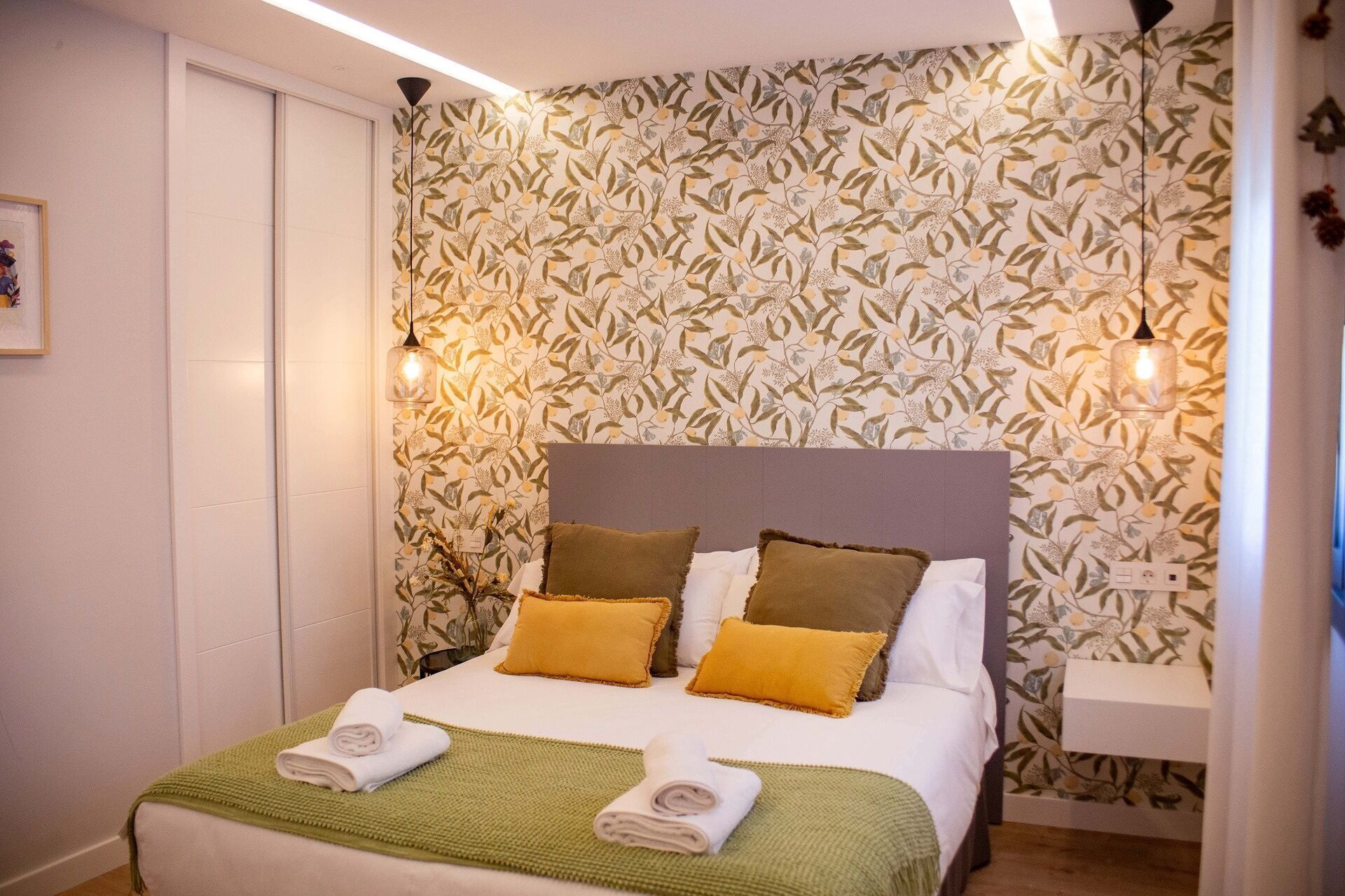 A neatly made bed with a green blanket and two pillows, a wall with a leafy pattern, and two wall-mounted lamps.