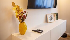 Une console blanche sur laquelle sont posés un vase jaune rempli de fleurs, deux télécommandes et un tableau encadré. Un téléviseur est accroché au mur au-dessus.