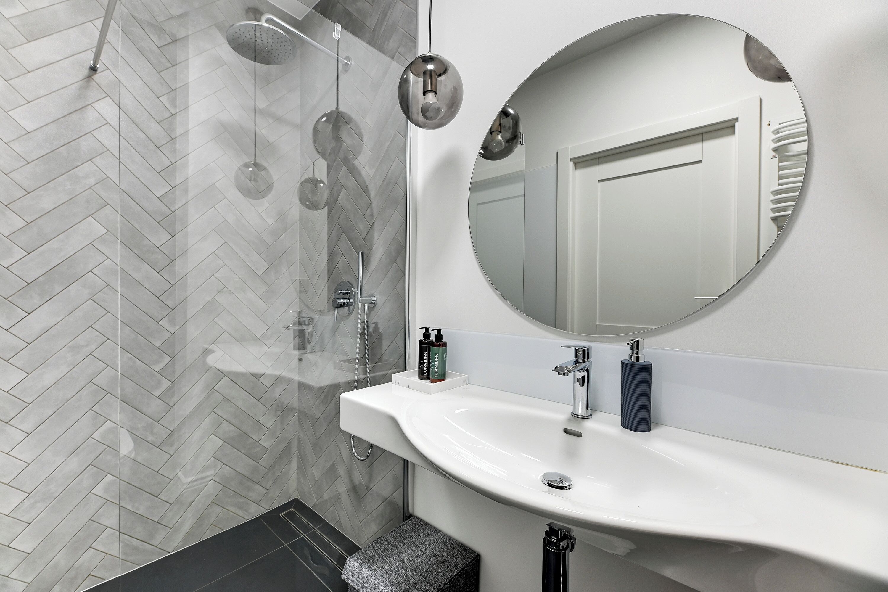 A modern bathroom with a herringbone tile floor, a large round mirror, a white sink, and a glass shower enclosure.