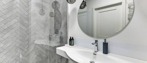 A modern bathroom with a herringbone tile floor, a large round mirror, a white sink, and a glass shower enclosure.