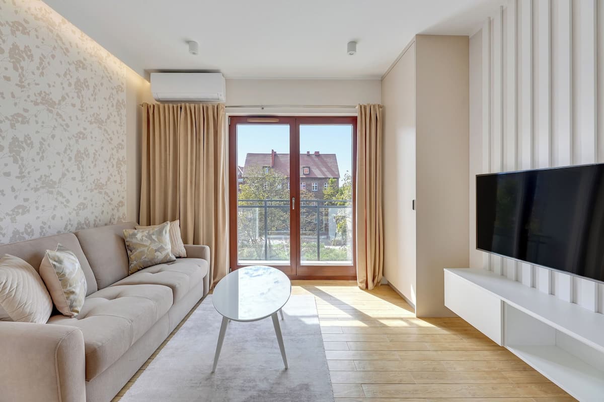Un moderno salón con sofá, una mesa de centro, televisor de pantalla plana y un balcón con vistas a edificios.