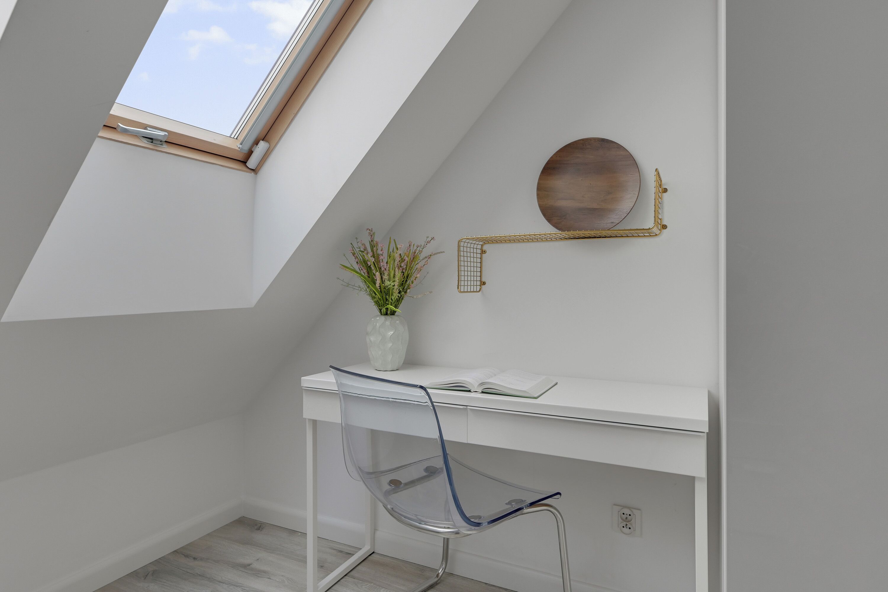 A modern, white room with a skylight, a desk with a vase of flowers, a chair, and a round mirror on the wall.