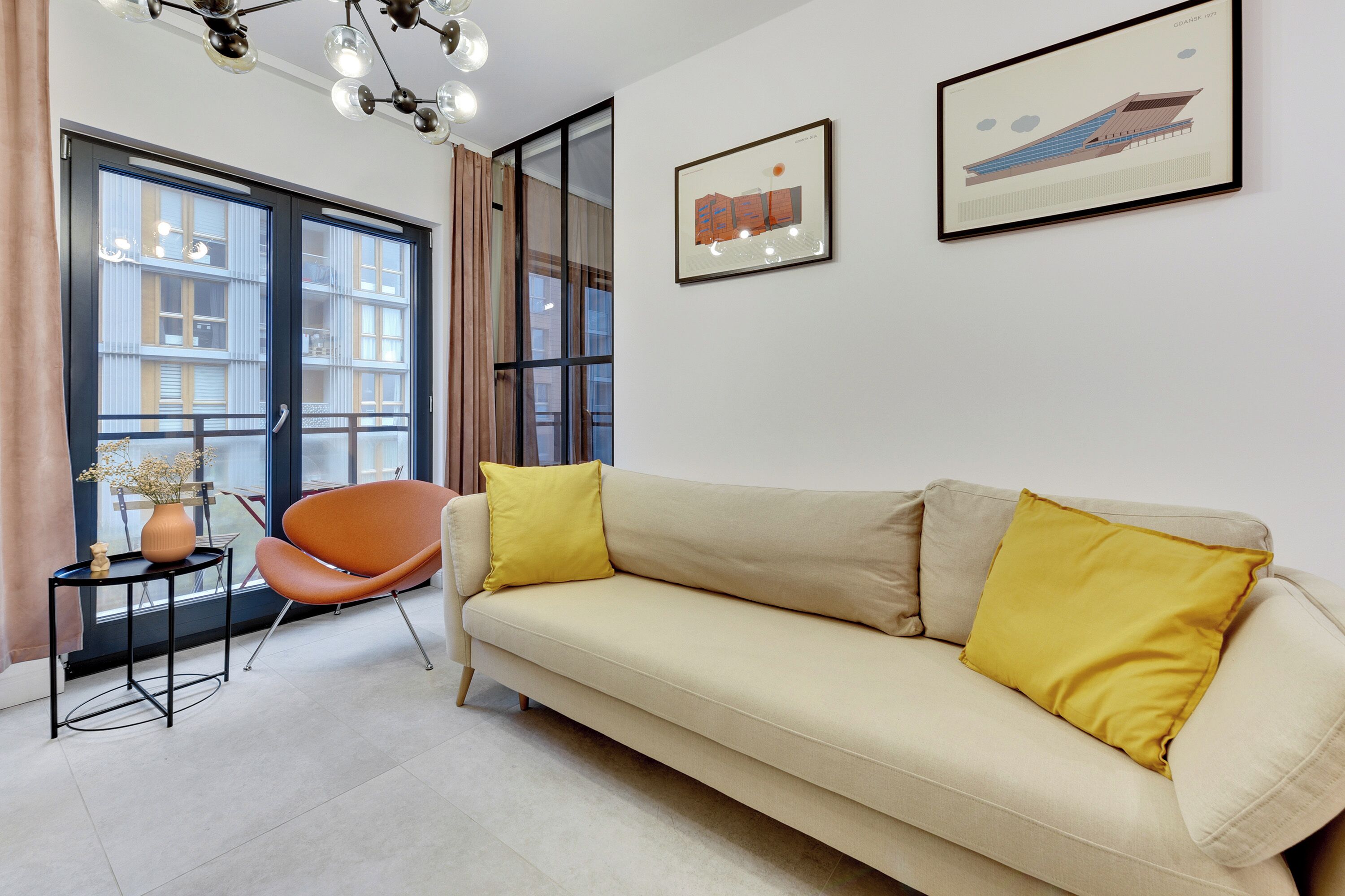 Un salon moderne avec un canapé beige, un coussin jaune, une table d’appoint noire et un vase rempli de fleurs. Il y a deux fenêtres avec des rideaux, un lustre et deux tableaux encadrés accrochés au mur.