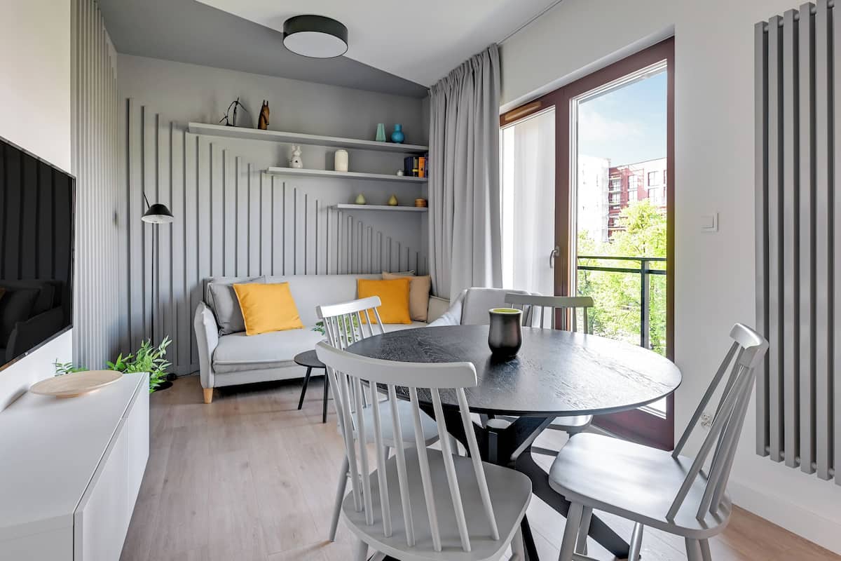 Una sala moderna con un sofá gris, sillas blancas, una mesa de comedor redonda y un balcón con vistas a edificios.