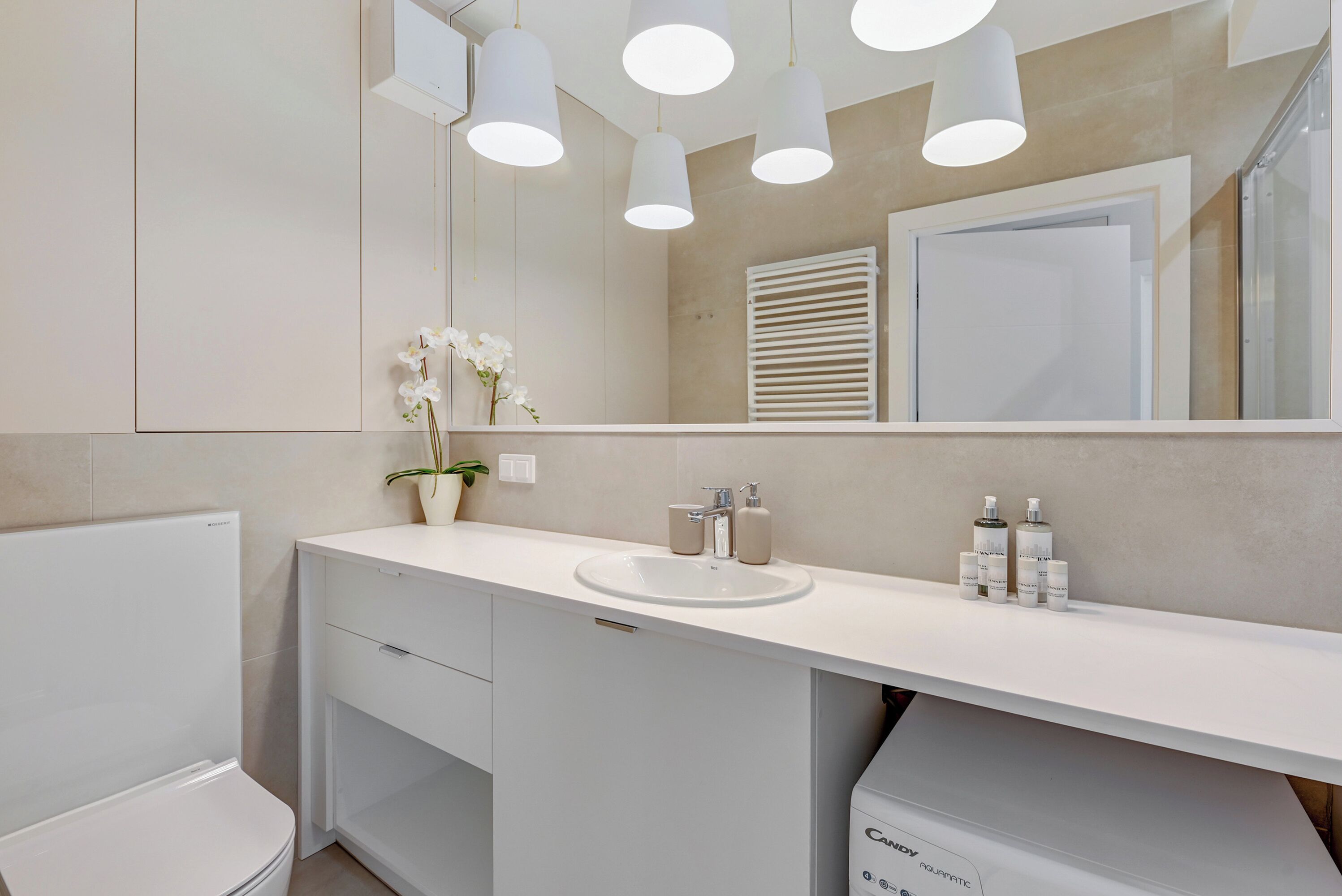 A modern bathroom with a white sink, mirror, and toilet. There are three pendant lights above the sink and a potted orchid on the counter.