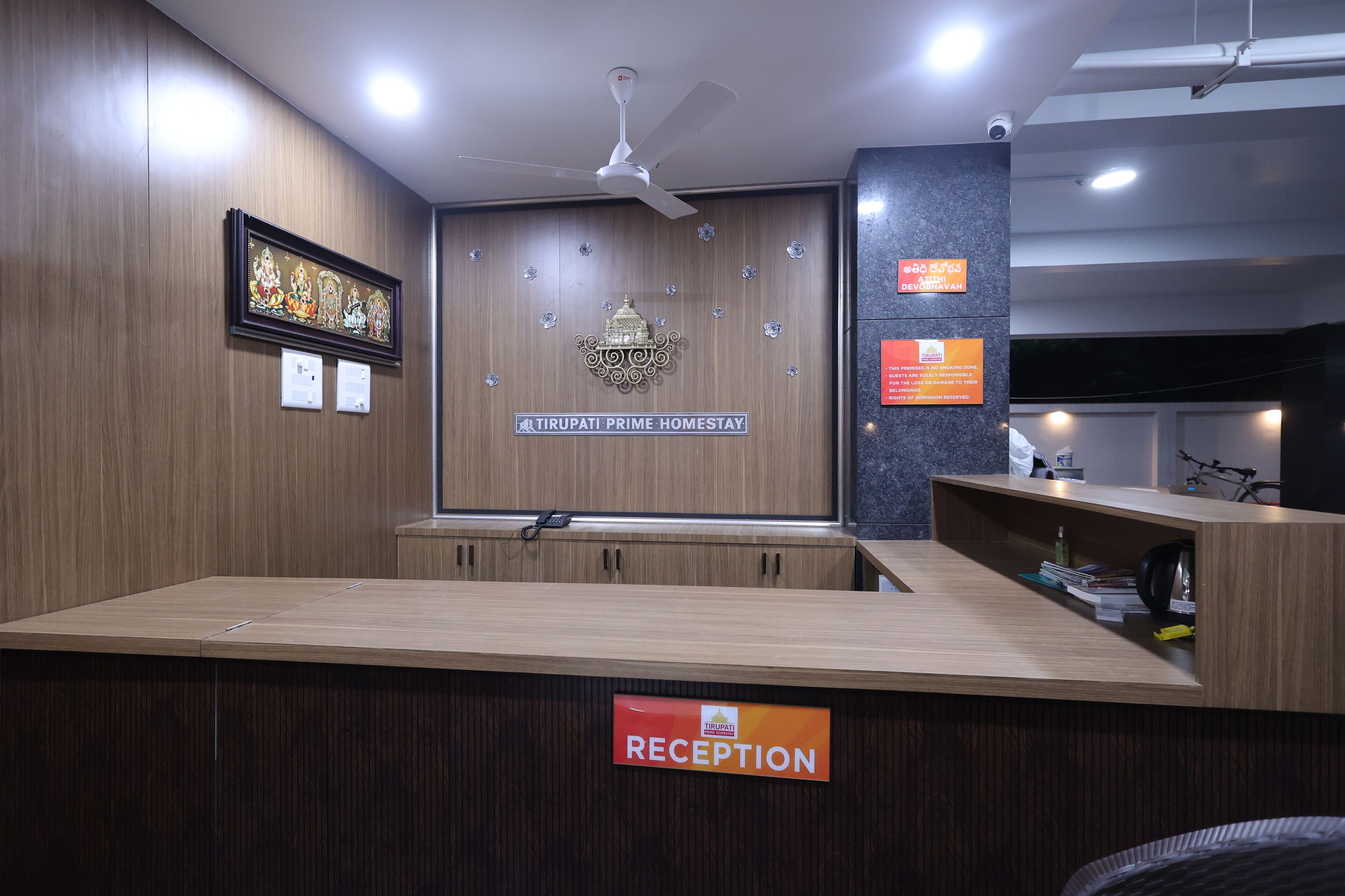 A reception area with a wooden counter, a ceiling fan, and a sign indicating 'Tirupati Prime Homestay'.