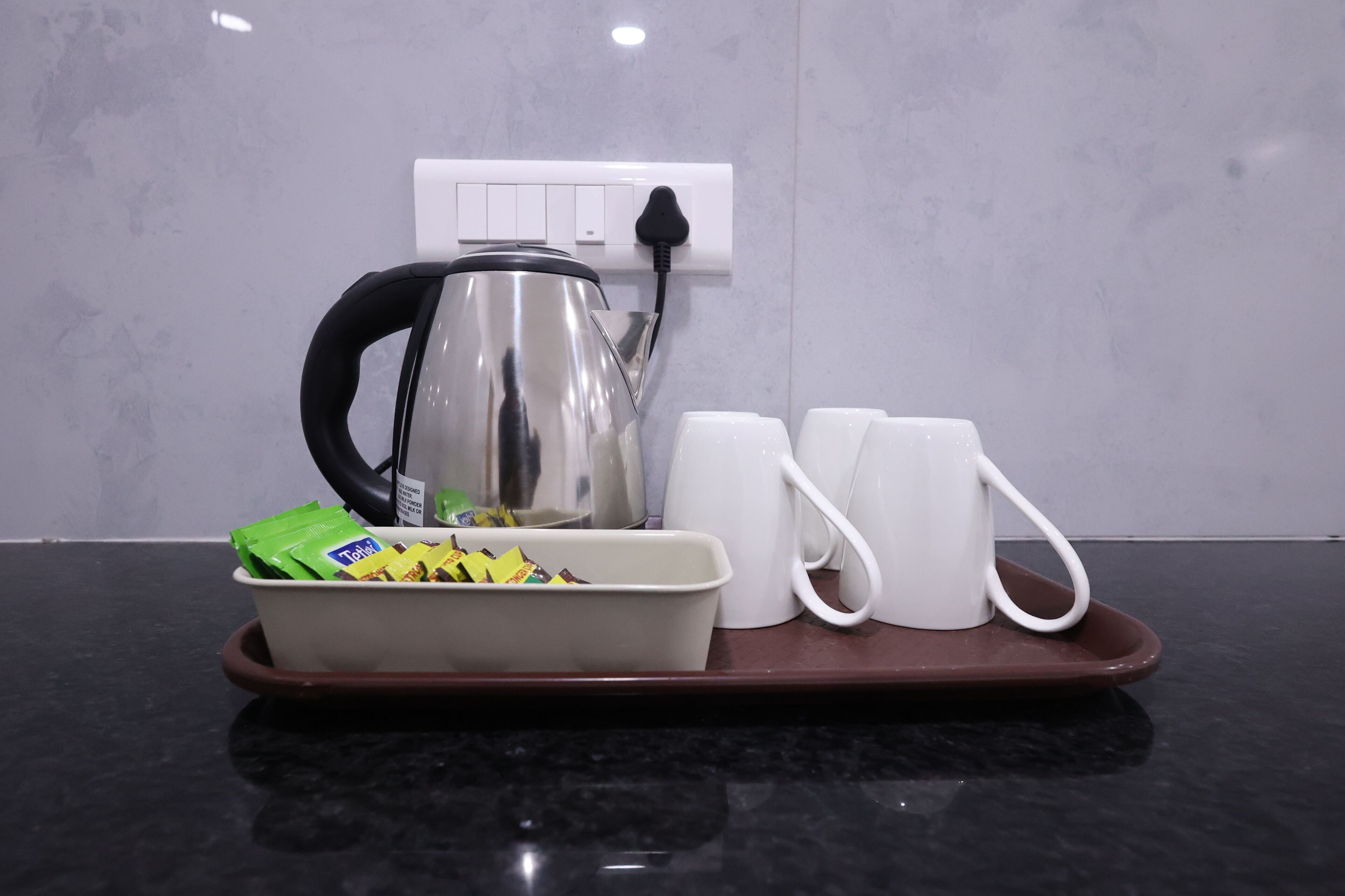 A tray with a kettle, two mugs, and a container of snacks on a black countertop.