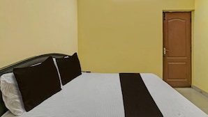 Una cama individual con colcha a rayas blancas y negras, cabecero negro y dos almohadas negras. La habitación tiene paredes amarillas y una puerta de madera.