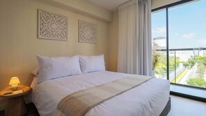 A hotel room with a large bed, two decorative wall panels, a nightstand with a lamp, and a view of a cityscape through a large window. - w* | Cozy 1BR w/ Balcony in San Isidro (Lima)