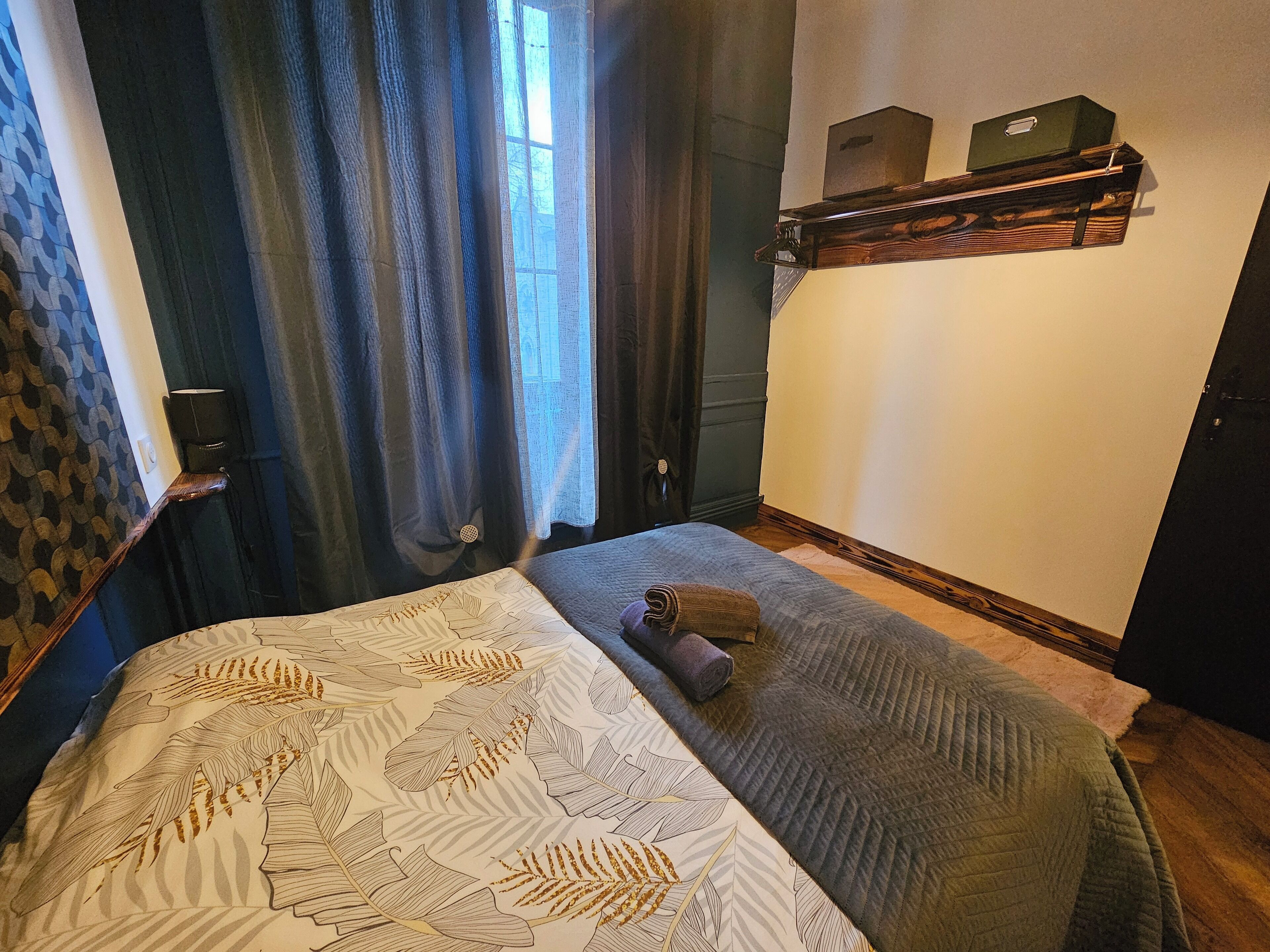 Una camera d'albergo con un letto, una finestra con tende, uno scaffale a parete con scatole e una coperta con motivo.