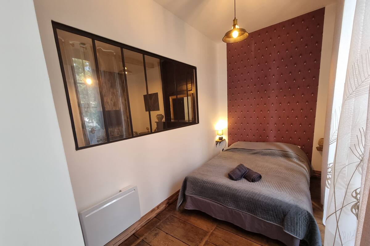 A bedroom with a bed, a large mirror, a red headboard, a wooden floor, and a window with a view of trees.
