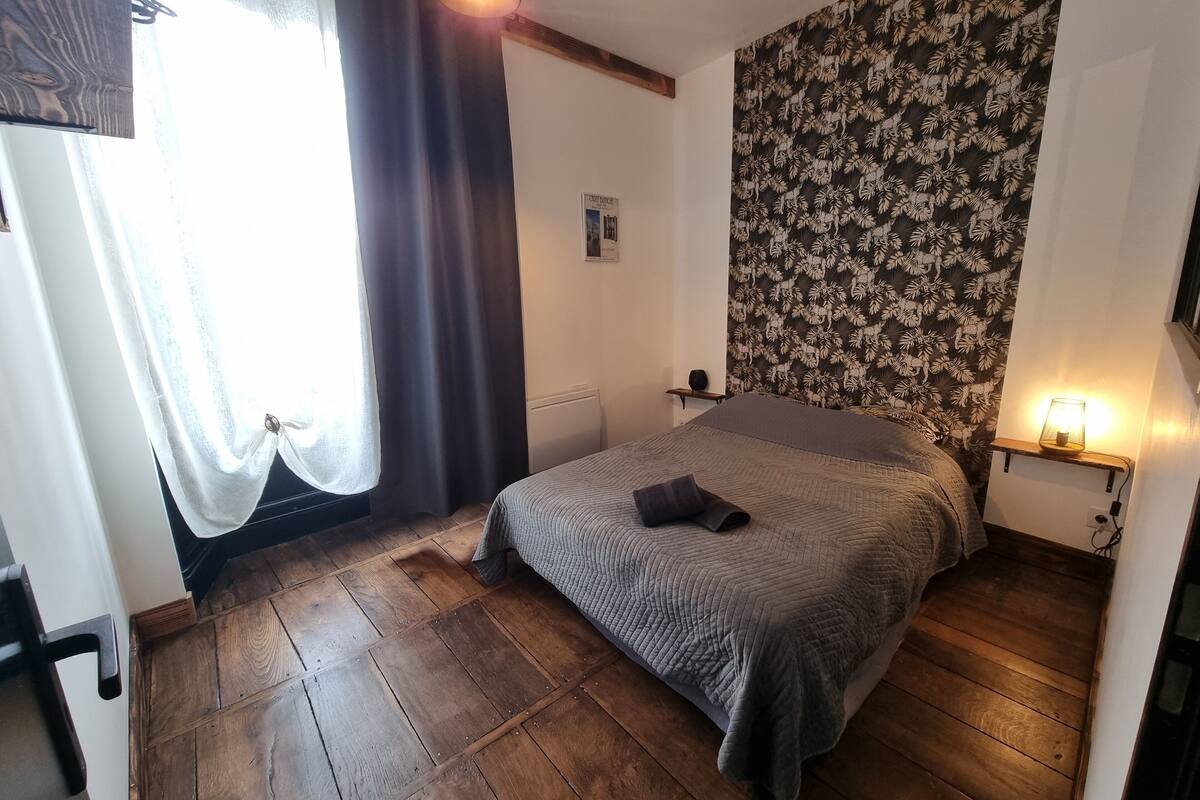 A bedroom with a wooden floor, a bed with a grey quilt, a nightstand with a lamp, and a floral wallpaper on the wall.