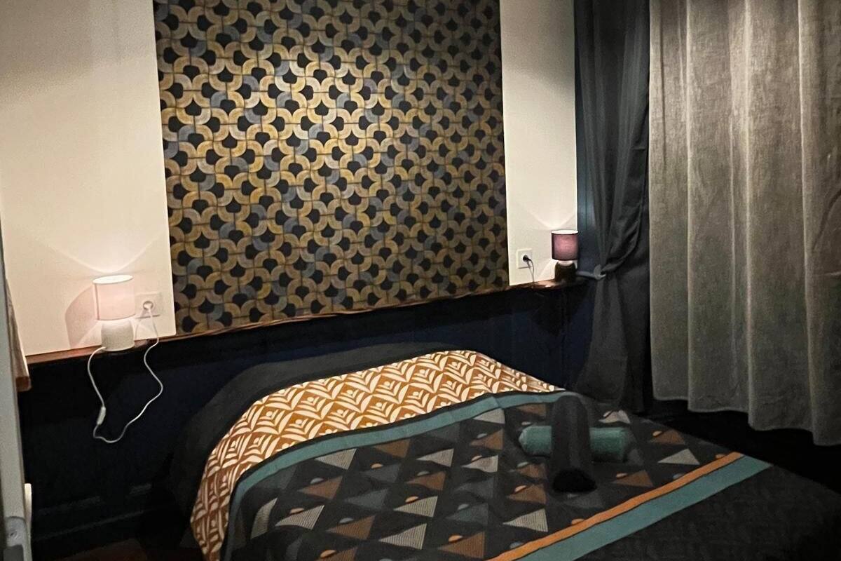 A room with a patterned wall, a bed with a geometric-patterned bedspread, a hanging light fixture, and a curtain.