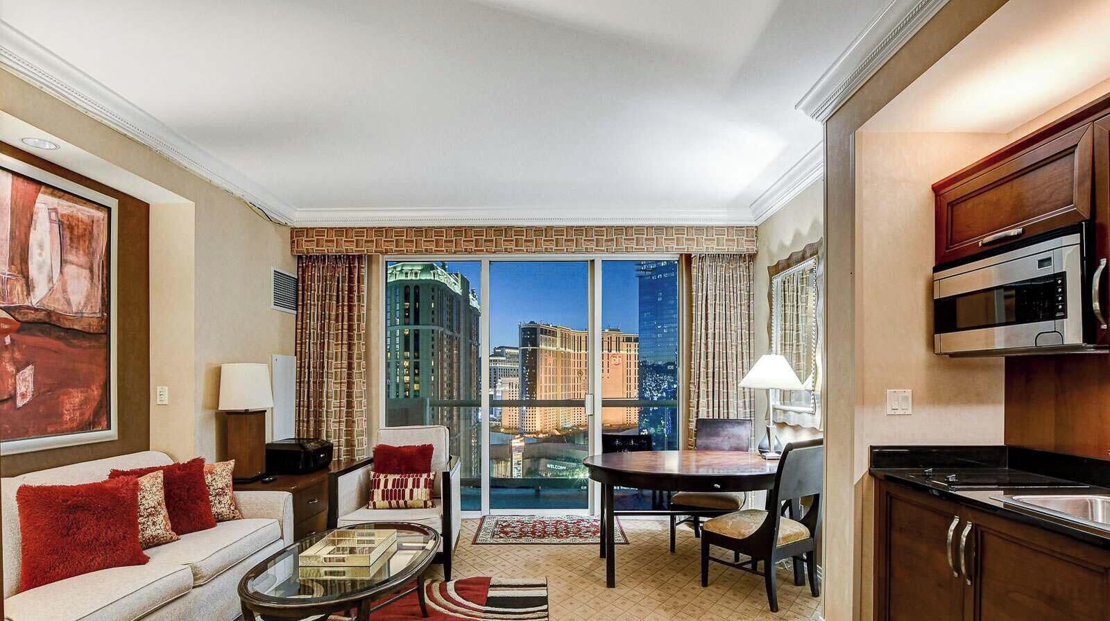 Quarto de hotel com varanda, área de jantar, sofá com almofadas vermelhas, mesa de centro, luminária e cozinha equipada com micro-ondas.