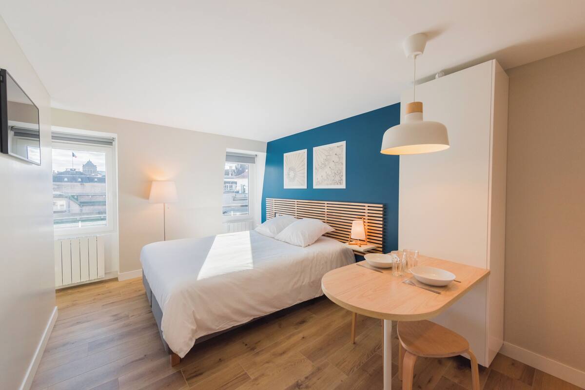 A bedroom with a bed, bedside table, lamp, and a view of the city through a window.