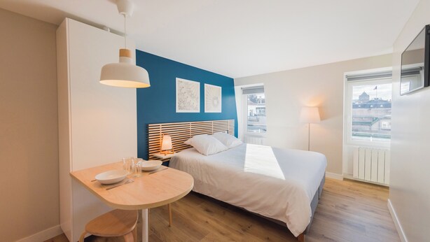 Un dormitorio con cama, mesita de noche y ventana con vistas a edificios.