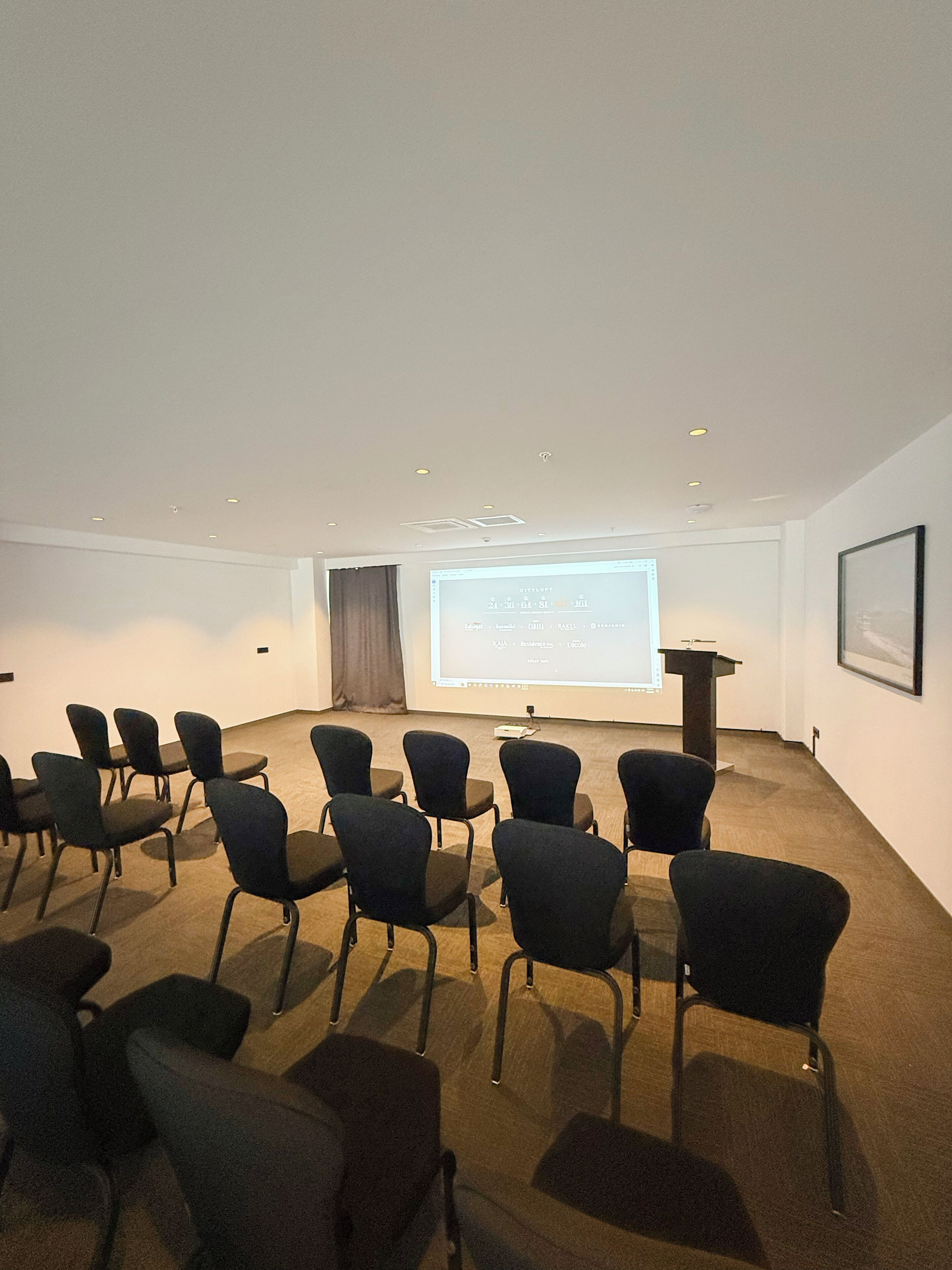A conference room with rows of chairs facing a projection screen, a podium, and a framed picture on the wall.