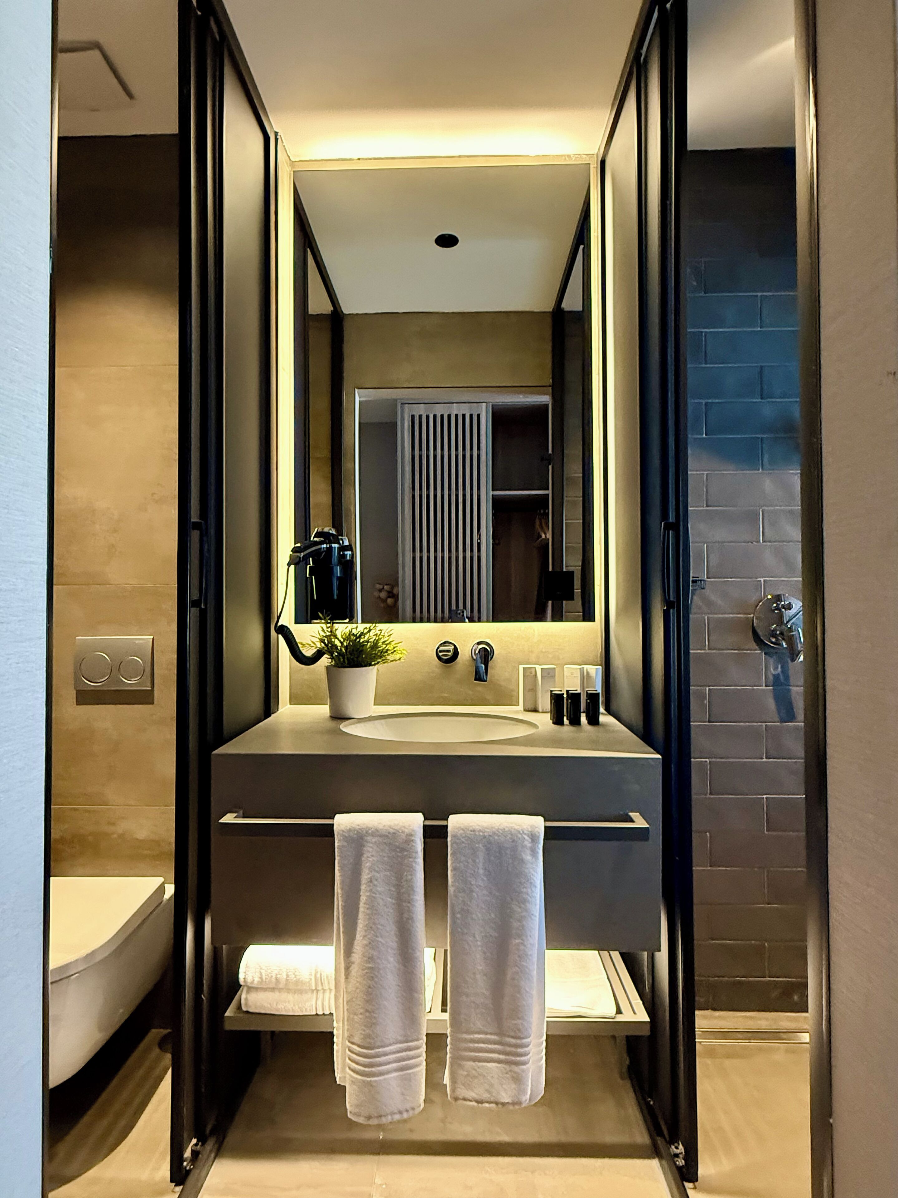 A modern bathroom with a large mirror, a sink, two towels, and a potted plant.