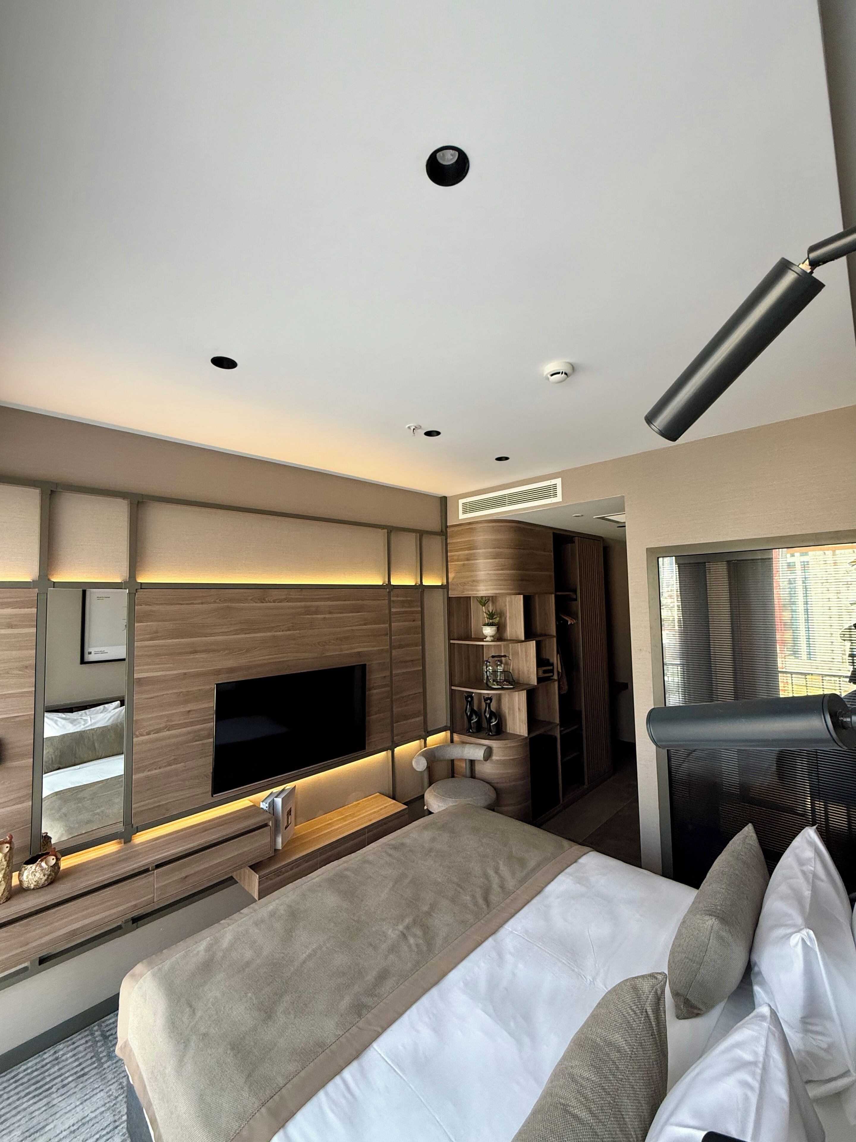 A modern hotel room with a large bed, a flat-screen TV, a built-in desk, and a large window with blinds.