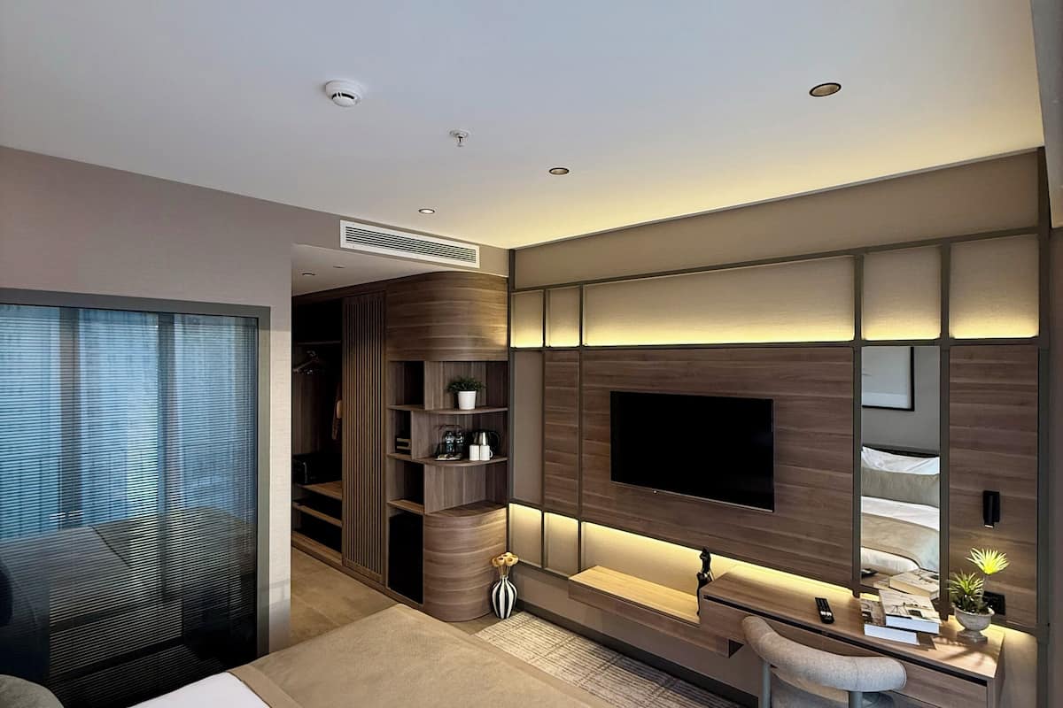 Ein modernes Hotelzimmer mit einem großen Bett, einem Schreibtisch mit Stuhl, einem an der Wand montierten Fernseher und einem begehbaren Kleiderschrank.