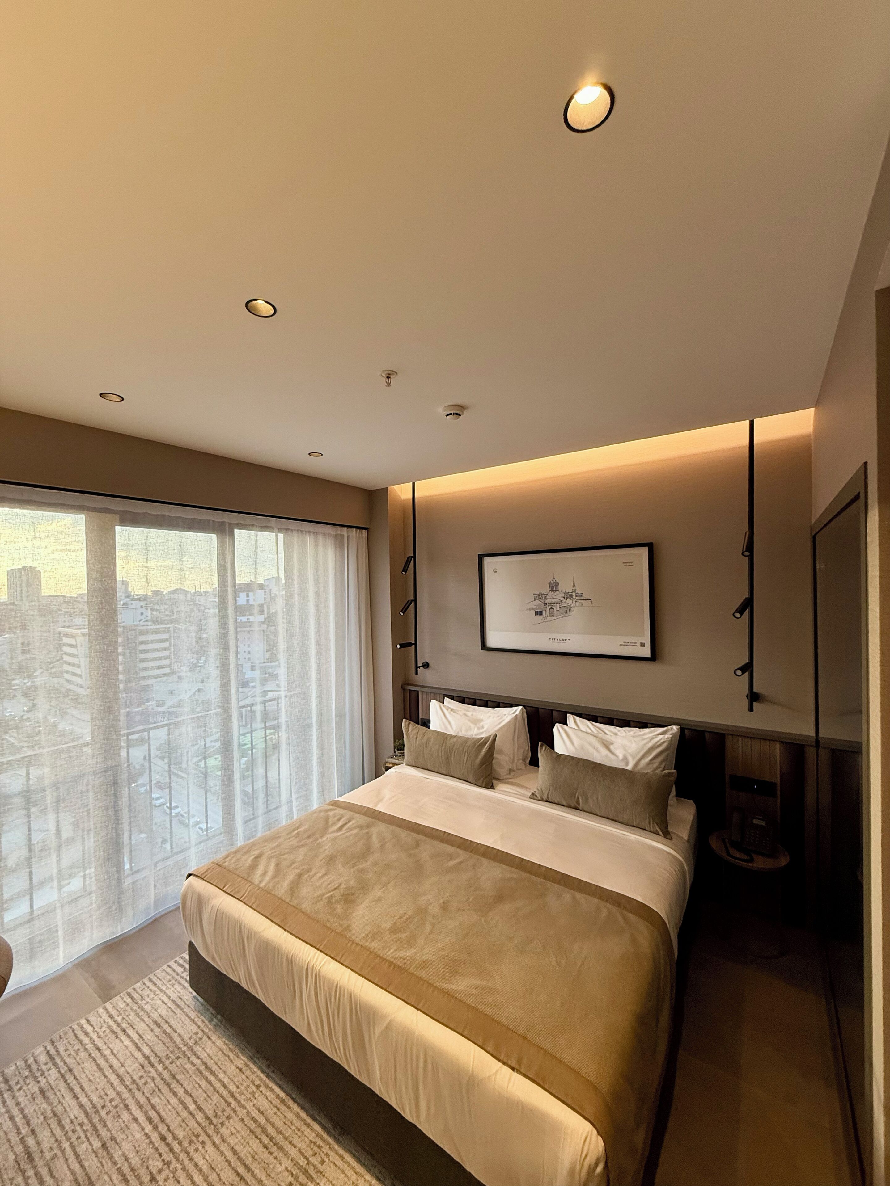 Un dormitorio de hotel moderno con una cama grande, vista a la ciudad y un cuadro enmarcado en la pared.