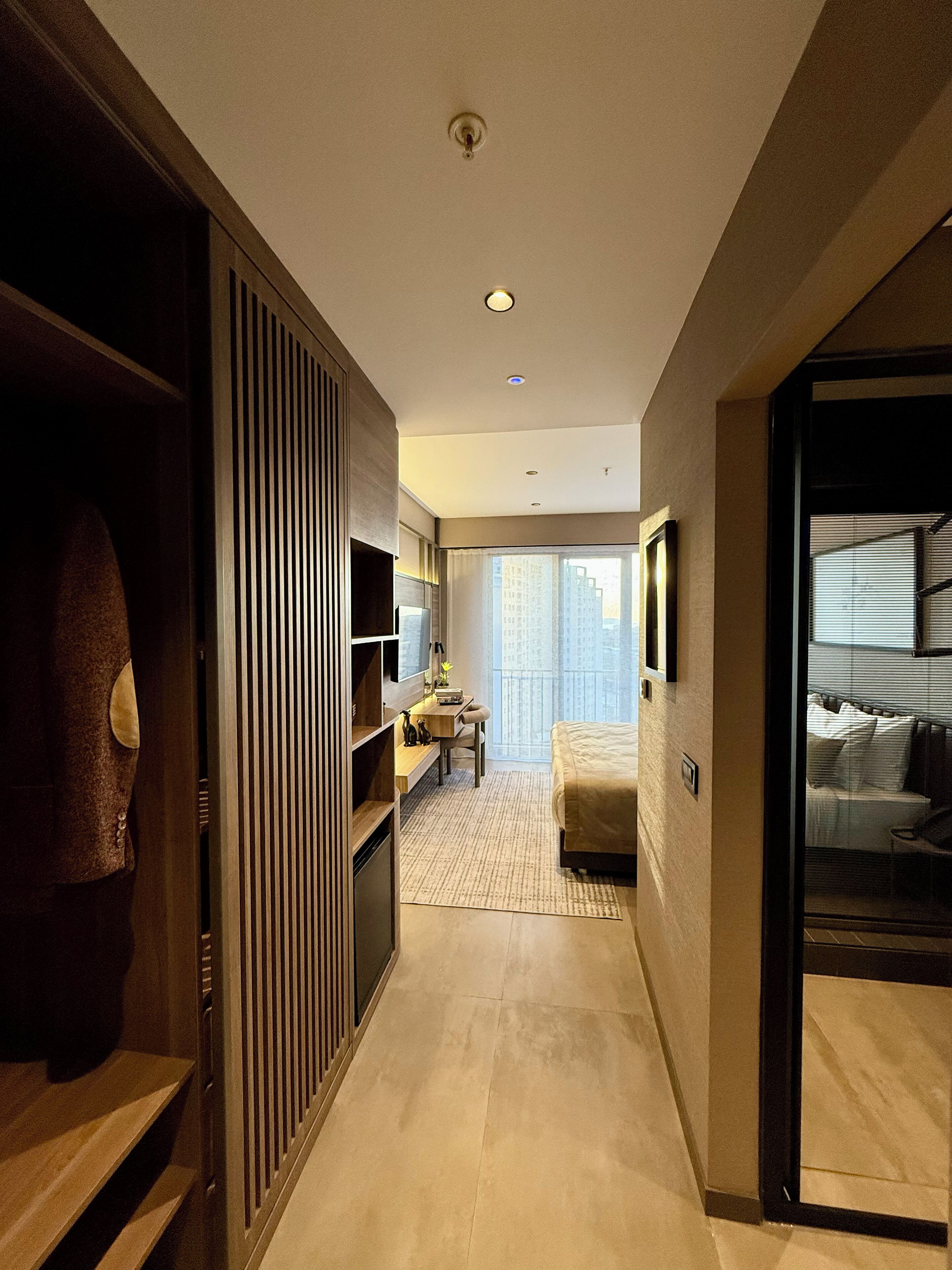 A hotel room with a bed, a desk, a wardrobe, and a view of the city.
