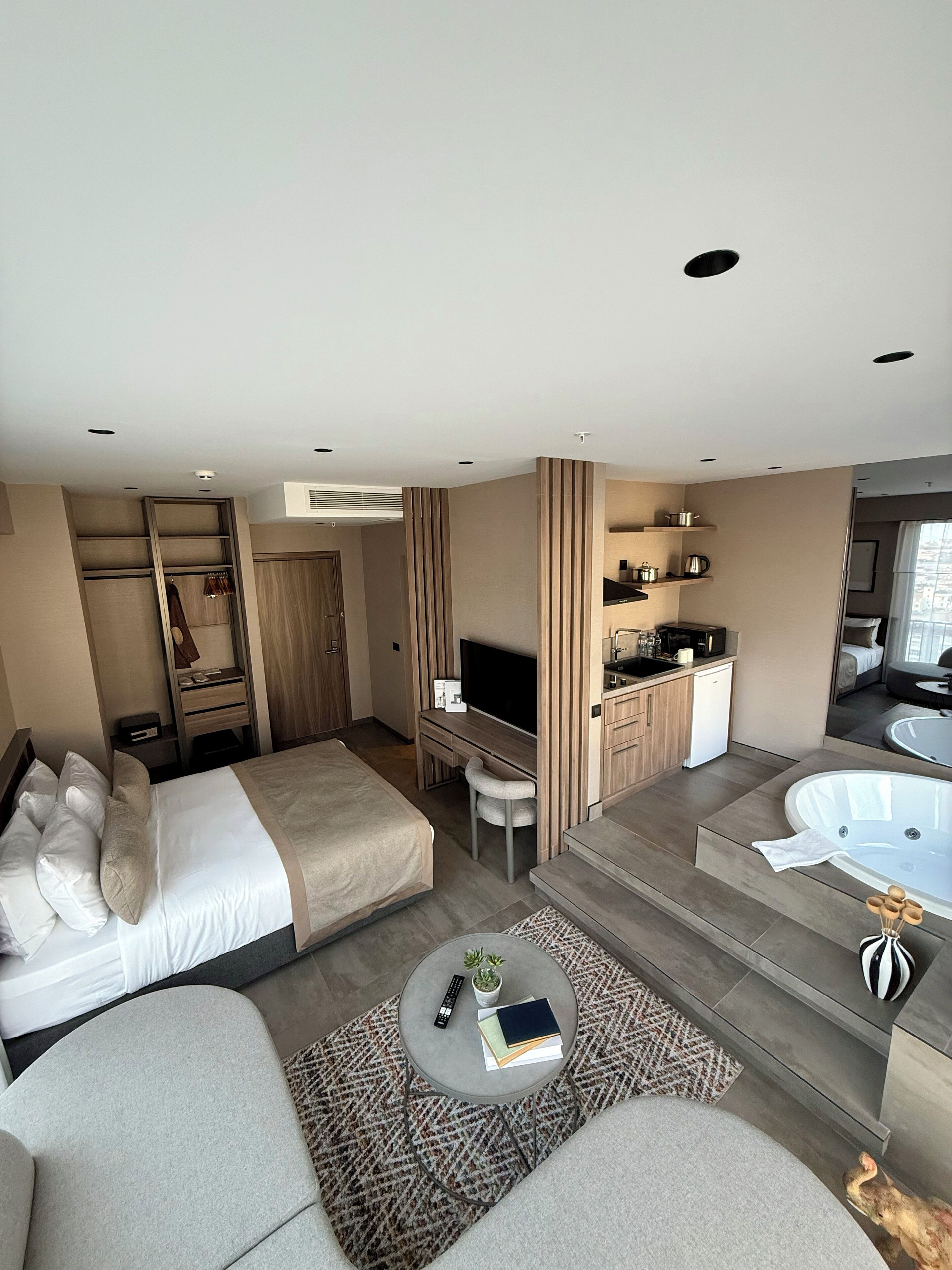 A modern hotel room with a bed, a sofa, a desk, a TV, a kitchen area, and a bathtub.