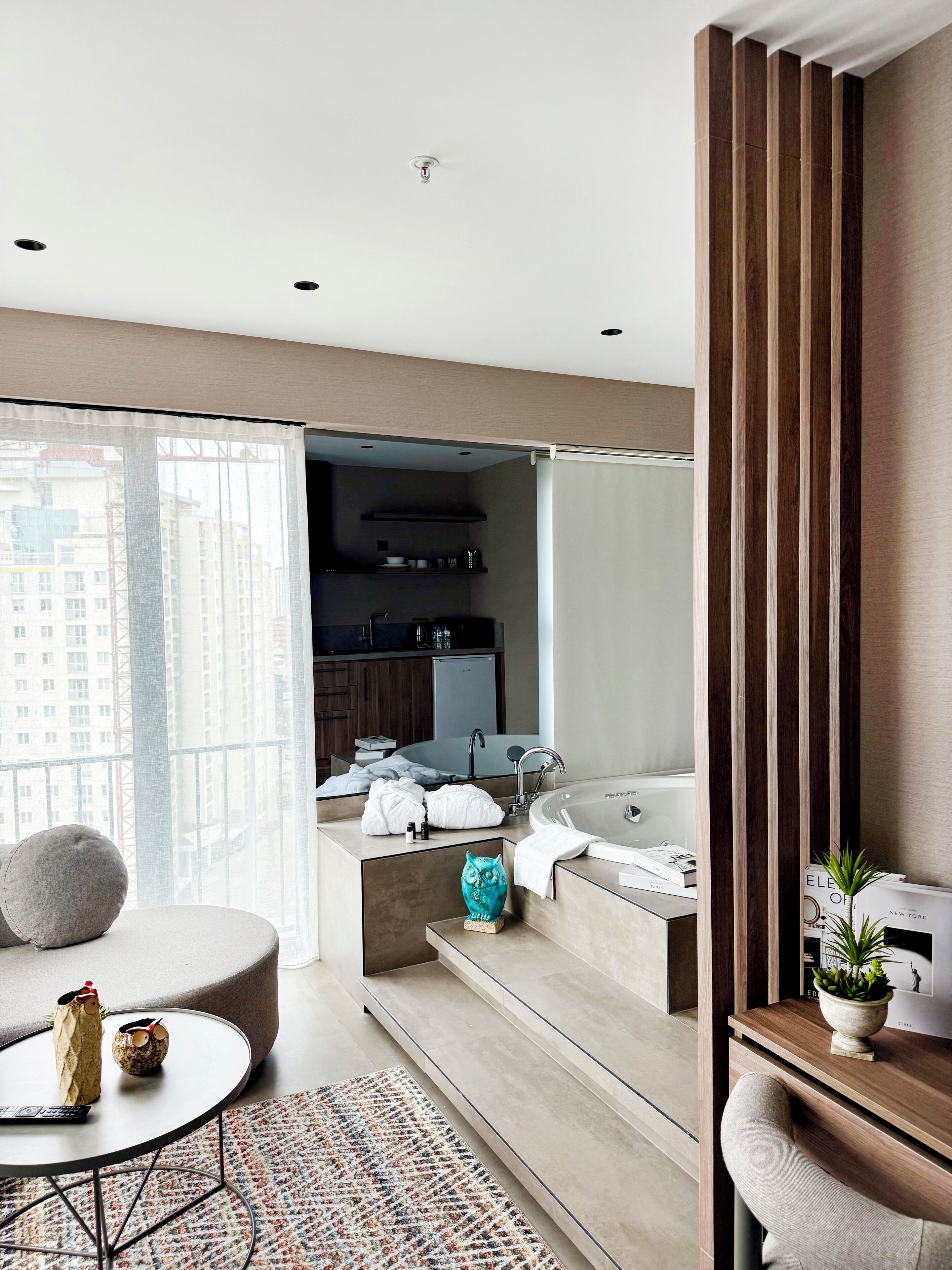 A modern hotel room with a balcony, a kitchenette, a large bathtub, and a seating area with a round table.
