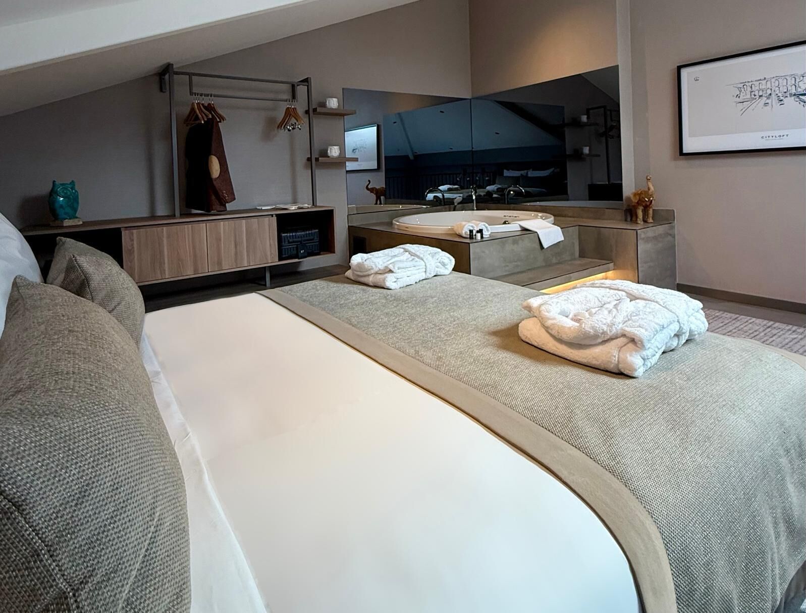 A modern hotel room with a large bed, a sitting area, a vanity with a mirror, and a bathtub.