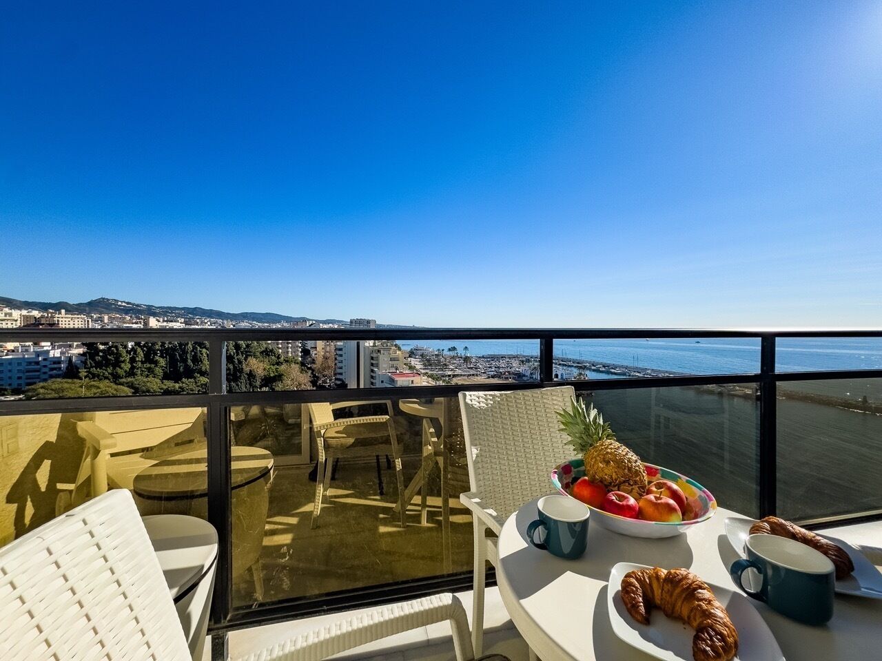 A balcony with a table set for breakfast, including fruits and pastries, overlooking a cityscape and the sea.
