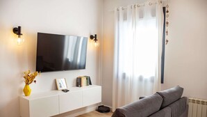Ein modernes Wohnzimmer mit einem Flachbildfernseher an einer weißen Wand, einem grauen Sofa, einem weißen Konsolentisch mit Büchern und einer Vase sowie einem Fenster mit transparenten Vorhängen.