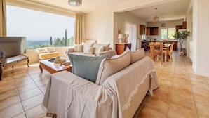 Un salon spacieux avec un canapé, une table basse et un téléviseur. Il y a également un coin repas avec une table et des chaises. La pièce est dotée de grandes fenêtres et offre une vue sur l’extérieur.