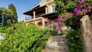 Un escalier en pierre mène à un bâtiment doté d’un balcon, le tout étant entouré d’une végétation luxuriante et de plantes en fleurs.