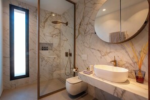 A modern bathroom with a marble wall, a large round mirror, a freestanding bathtub, and a toilet.