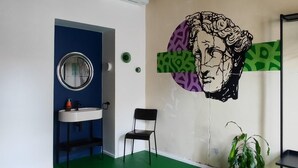 Une pièce avec un sol vert, un miroir rond, un lavabo, une chaise, une plante en pot et un mur orné d’un motif graphique.