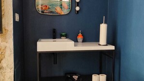 Une salle de bain avec un lavabo blanc, un miroir et des toilettes. On trouve des articles de toilette sur le lavabo et une poubelle à côté des toilettes.