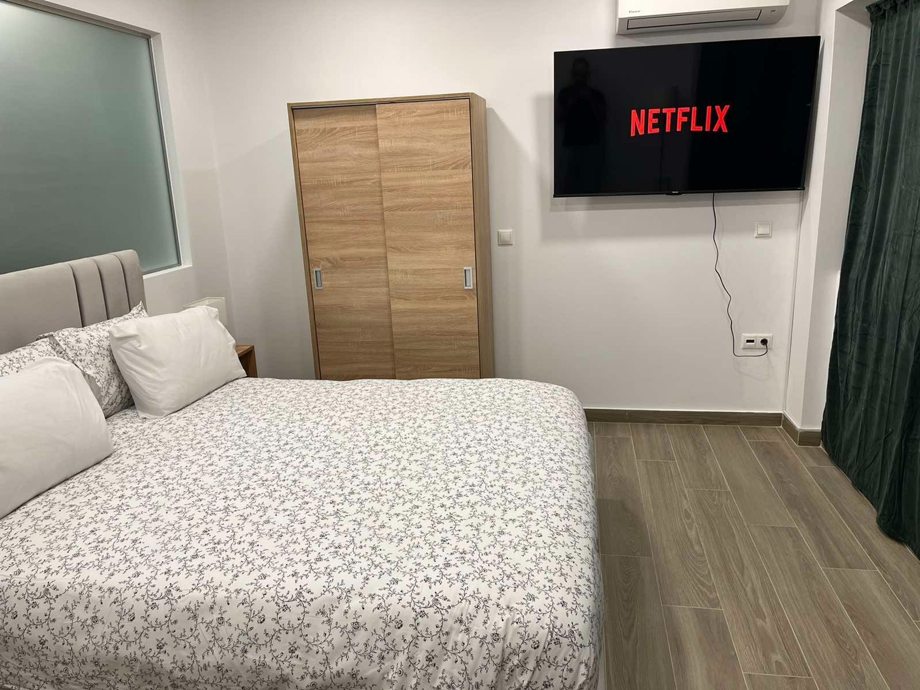A bedroom with a bed, a wardrobe, a wall-mounted TV displaying 'NETFLIX', and a window.
