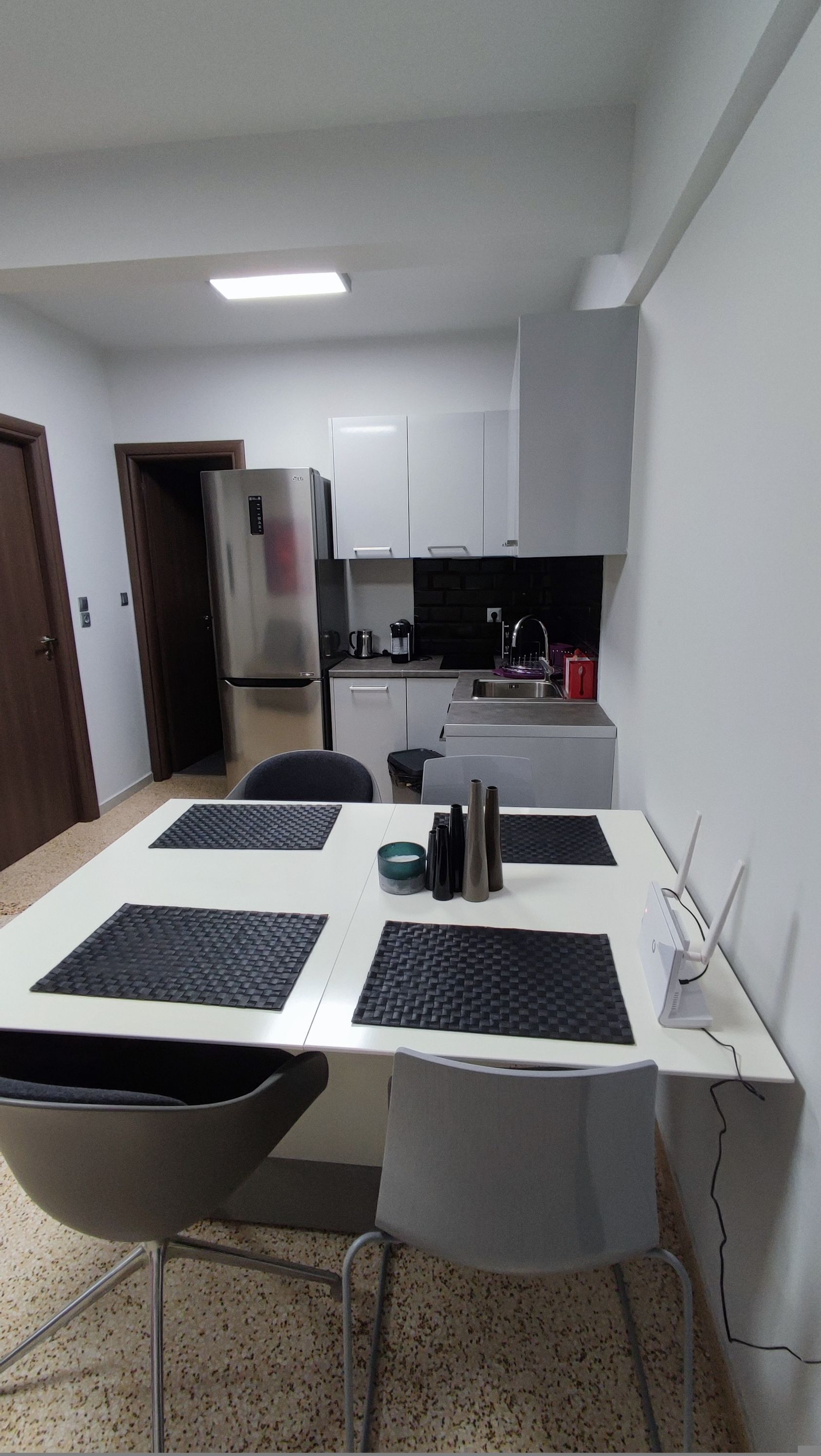 Una cocina compacta con armarios blancos, un refrigerador de acero inoxidable y una zona de comedor con mesa y sillas.