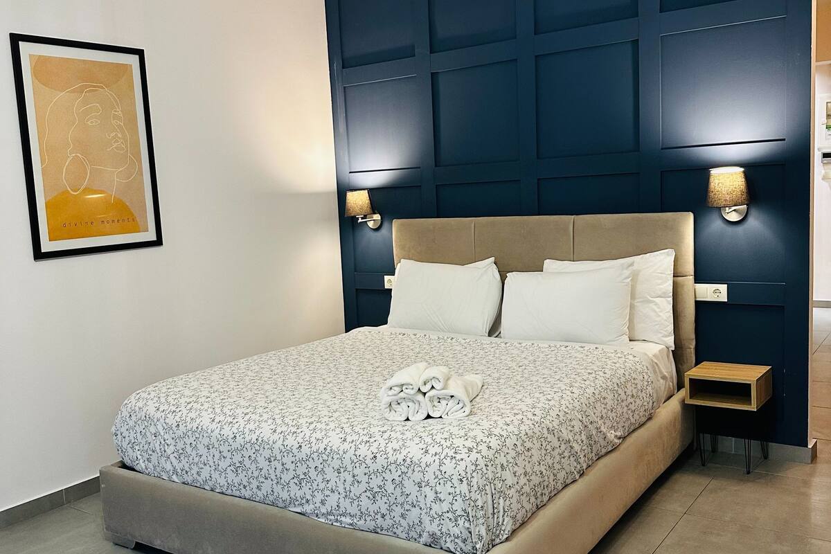 Ein ordentlich bezogenes Bett mit floralem Überwurf, ein Kopfteil mit integrierter Beleuchtung und ein gerahmtes Kunstwerk an der Wand.