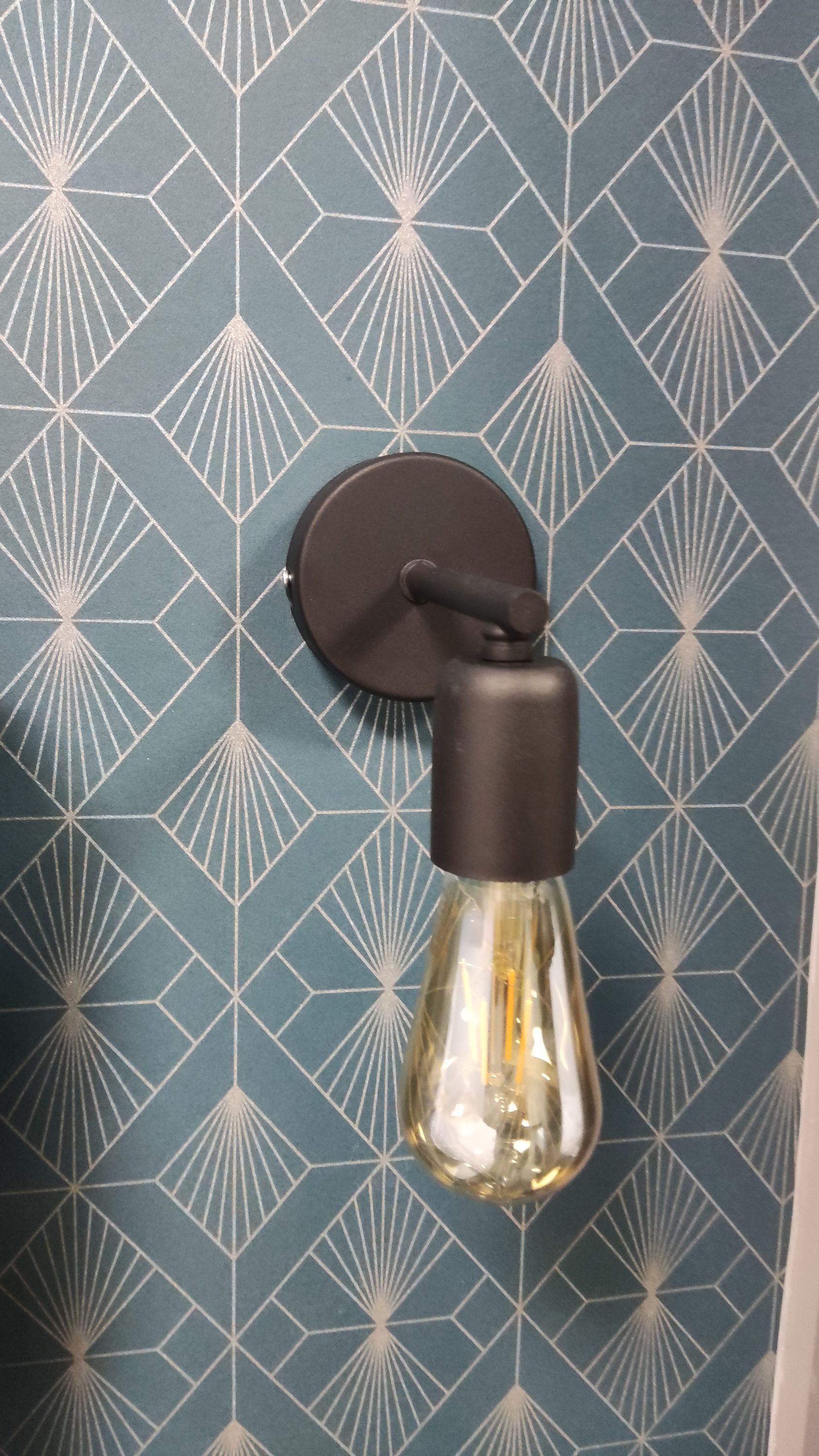 A wall-mounted light fixture with a round base and a glass enclosure, set against a patterned wallpaper with geometric shapes.