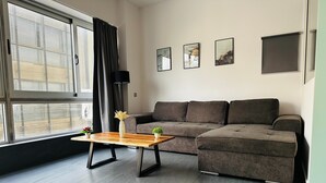 Ein modernes Wohnzimmer mit einem Ecksofa, einem Couchtisch aus Holz mit Pflanzen und einem großen Fenster mit Jalousien.