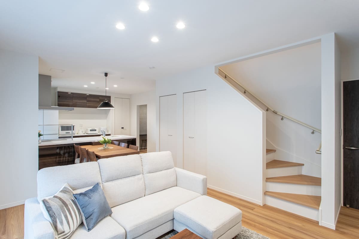 A modern living room with a white sofa, a dining area, and a staircase.