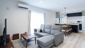 A modern living room with a grey sectional sofa, a wooden coffee table, and a TV stand. A kitchen with white cabinets and a dining area with a wooden table and chairs is visible in the background. - MOVA Villa Ozu (Ozu)