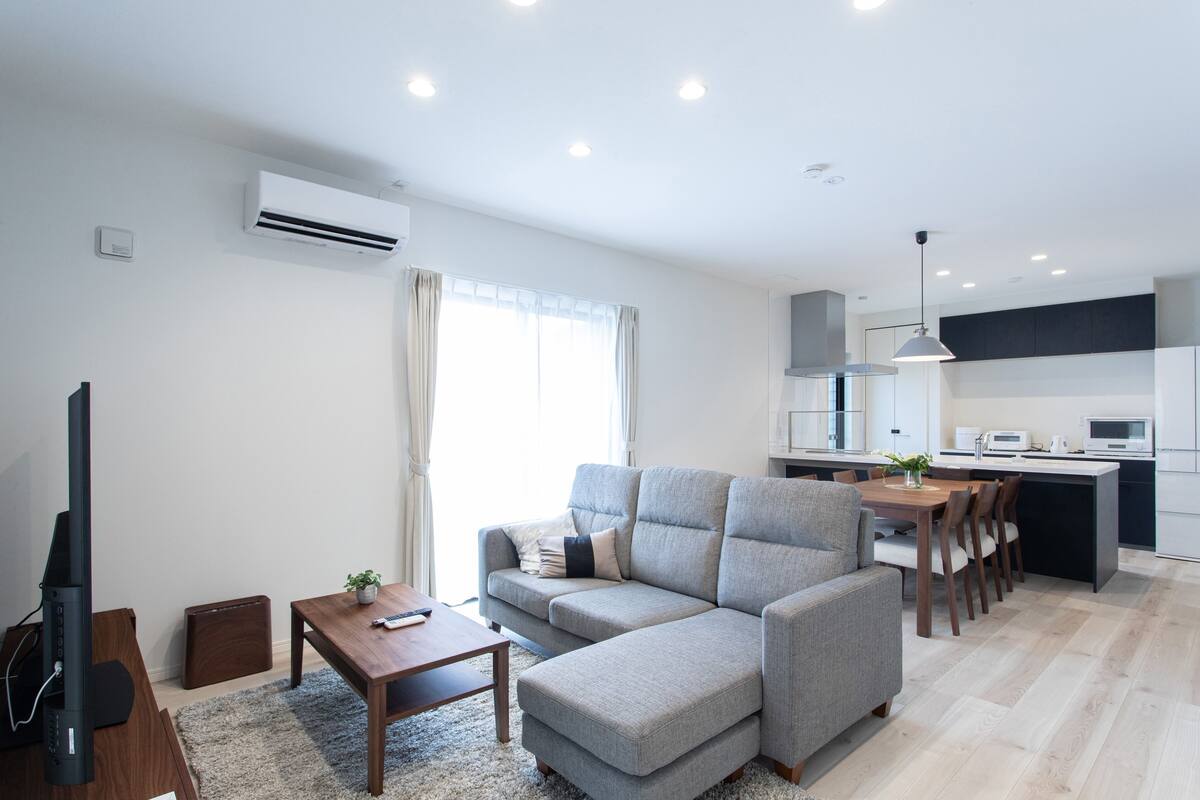 A modern living room with a grey sectional sofa, a wooden coffee table, and a TV stand. A kitchen with white cabinets and a dining area with a wooden table and chairs is visible in the background.