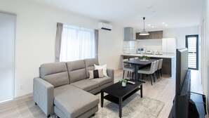 A modern living room with a sofa, coffee table, and TV. There is a dining area with a table and chairs. The kitchen is visible in the background with cabinets and appliances. - MOVA Villa Ozu (Ozu)